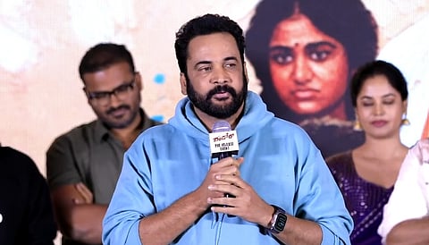 A medium shot of Telugu actor Sivaji speaking into a microphone at a pre-release event for the film Dhandoraa.

He is dressed in a light blue hooded sweatshirt and has a full black beard. He holds a microphone that has a white "Dhandoraa Pre-release Event" label on it. In the background, there is a large poster featuring a woman's face and several other people standing behind him, including a man in a green shirt and a woman in a purple saree.