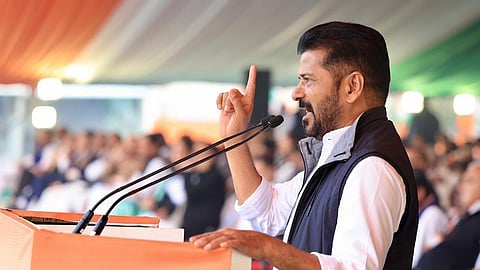 A medium shot from a side profile captures Revanth Reddy, the Chief Minister of Telangana, speaking passionately at an outdoor political rally. He is a middle-aged Indian man with short, dark hair and a well-groomed beard and mustache. He wears a crisp white long-sleeved shirt with the sleeves rolled up to his elbows, topped with a dark navy blue sleeveless quilted vest.

Standing behind a bright orange podium, he leans slightly forward with his left hand resting on the edge. His right arm is raised, with his index finger pointing upward in an assertive, emphatic gesture. Two microphones on long, thin stands are positioned in front of him.

The background shows a large, blurred crowd of people seated under a massive tent or canopy. The canopy features stripes of orange, white, and green, suggesting the colors of the Indian national flag. The lighting is bright and natural, indicating an afternoon event, creating a focused and energetic atmosphere.