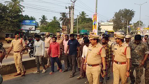 An eye-level shot shows several people walking down a road in East Godavari, Andhra Pradesh, India. In the center, several men in plain clothes are walking with their hands behind their backs, escorted by many police officers in khaki uniforms and one officer in camouflage. The police officers are walking alongside and behind the men, and some are holding sticks. The road is paved and there are several vehicles and people in the background. On the left side of the road, there is a low concrete wall and some trees. On the right side of the road, there are some buildings and power lines. The sky is clear and the sun is shining. In the background, there are posters on poles. The man on the far left is wearing a green shirt and is partially visible. The man next to him is wearing a grey shirt and jeans. The man next to him is wearing a red shirt and blue pants. The man next to him is wearing a pink shirt and grey pants. The man next to him is wearing a navy blue shirt and grey pants. The man next to him is wearing a brown shirt and grey pants. The man next to him is wearing a yellow and black plaid shirt and blue jeans. The police officers are wearing khaki uniforms with brown belts and hats. One officer is wearing a camouflage uniform. The background shows a busy street with people, motorcycles, and buildings. The lighting is bright and even, suggesting it is daytime.