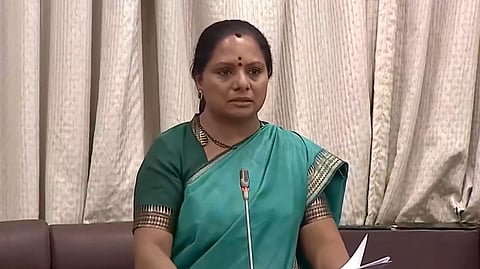 A medium shot of K. Kavitha, an Indian politician, standing behind a microphone in what appears to be a legislative assembly or council chamber. She is wearing a teal green saree with a gold-patterned border and a matching green blouse. She has a bindi on her forehead and is looking forward with a serious expression, holding papers in her hand. The background consists of light-colored, pleated vertical curtains and a dark brown seating area.