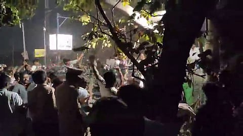 A low-angle, nighttime shot captured from a slightly elevated position behind a tree, showing a large, crowded gathering outdoors. In the foreground, the dark, textured bark and green leaves of a tree frame the right side of the image. The crowd is composed of many people, some with their backs to the camera, and others facing toward a bright, glowing square light or billboard in the background. Several individuals have their arms raised. The scene is illuminated by the bright light and overhead street lamps, which create a hazy, misty effect in the air. A person in a uniform, a police officer, is visible in the lower-middle section of the crowd. The overall mood is somewhat chaotic.