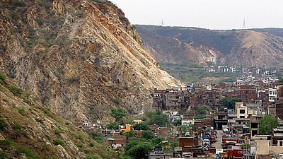 A densely packed residential area nestled at the base of a steep, rugged mountain in the Aravalli Range near Jaipur, India. The mountain features prominent geological layers of light brown and tan rock, with sparse green vegetation clinging to its slopes. The urban settlement consists of multi-story brick and concrete buildings crowded together, with a few trucks visible on a narrow street, illustrating the close proximity between the natural landscape and urban expansion. In the background, rolling hills continue under a hazy, overcast sky.