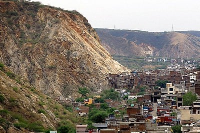 A densely packed residential area nestled at the base of a steep, rugged mountain in the Aravalli Range near Jaipur, India. The mountain features prominent geological layers of light brown and tan rock, with sparse green vegetation clinging to its slopes. The urban settlement consists of multi-story brick and concrete buildings crowded together, with a few trucks visible on a narrow street, illustrating the close proximity between the natural landscape and urban expansion. In the background, rolling hills continue under a hazy, overcast sky.