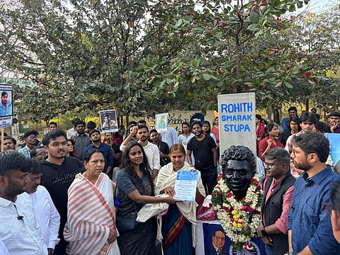 A medium-shot, outdoor photograph shows a group of people gathered around a memorial for Rohith Vemula. In the center, Radhika Vemula, wearing a white shawl, stands next to a woman in a dark patterned dress; together, they hold up a blue and white document titled "Model Legislation: Karnataka Rohith Vemula Act, 2024."