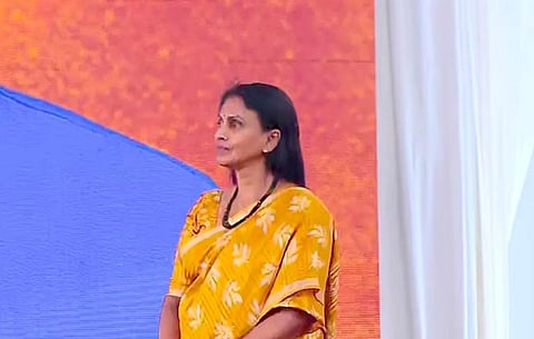 Sreelekha in a yellow saree stands on a stage, looking to the side, with a large orange and blue backdrop behind her. White drapes frame the stage, suggesting a formal or public event setting