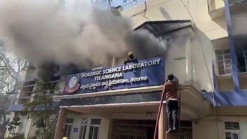 A wide, medium shot shows dark grey smoke billowing from the second-story windows of the Forensic Science Laboratory building in Telangana.

The building is a multi-story, cream-colored structure with blue trim. A large blue sign above the main entrance clearly identifies the facility in both English and Telugu. In the foreground, a firefighter wearing a yellow helmet is visible on a balcony near the smoke, while another person in a brown shirt climbs a ladder leaning against the building’s entrance overhang. The scene captures an active emergency response to a structural fire.