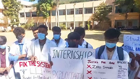 Young men stand in a row wearing white lab coats and wearing masks, holding up hand made posters with slogans such as 'Fix the System', 'Internship not exploitation', and 'Justice for AHS'. Behind them is long building with columns running on the outer wall. 