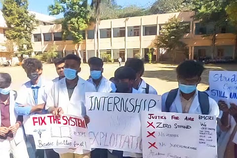 Young men stand in a row wearing white lab coats and wearing masks, holding up hand made posters with slogans such as 'Fix the System', 'Internship not exploitation', and 'Justice for AHS'. Behind them is long building with columns running on the outer wall. 