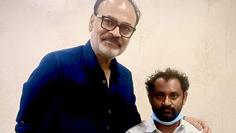 A medium shot captures two men standing indoors against a light-colored, patterned wall.

The man on the left is tall, wearing a dark blue button-up shirt and black-rimmed glasses. He has a salt-and-pepper beard and is smiling slightly at the camera with his hand resting on the other man's shoulder.

The man on the right is shorter, wearing a light-colored, long-sleeved shirt with a faint leaf pattern. He has dark hair and a beard, and is wearing white disposable gloves and a blue surgical mask tucked under his chin. He is looking directly at the camera with a neutral expression.

The lighting is bright and even, and the background features a subtle wallpaper with a faint tree branch motif.