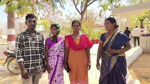 Four people, one man and three women, stand outdoors in a line under the shade of trees. The man on the left wears a black and white checkered shirt. The woman next to him wears a purple and white patterned sari with a green and black striped head covering. The woman in the center wears an orange tunic with a bright pink dupatta. The woman on the right wears a dark blue sari with a gold border. In the background, there are trees, a white building, and a few other people. The ground is sandy, and a motorcycle is partially visible on the left.