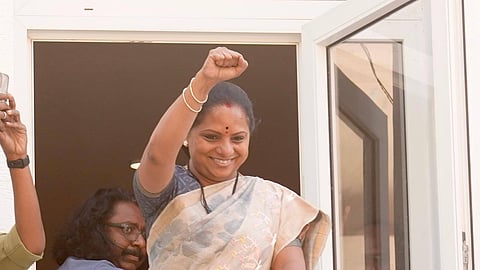 A medium-length, eye-level shot shows Kalvakuntla Kavitha, an Indian politician, standing on a balcony with a glass railing. She is smiling and has her right arm raised with a clenched fist in a gesture of victory or solidarity. She is wearing a cream-colored saree with a subtle floral pattern and a grey blouse, accessorized with gold bangles and a red bindi. Behind her to the left, a man with long dark hair and glasses is partially visible, and another person's arm holding a phone is seen on the far left edge. The background shows a white door frame and a glimpse of an interior room.