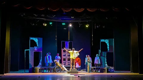 A scene from 'Bhagavantana Marana'. One actor in a yellow shirt and white scarf draped around hi sneck is standing in the middle atop a low stool, with his arms stretched out. Others form a semi circle behind him and look on