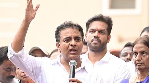 An Indian man, identified as politician K. T. Rama Rao (KTR), stands in the center of a crowd, speaking passionately into a handheld microphone. He is wearing a white button-down shirt and has his right arm raised high in a gesturing motion.

Directly behind him stands another man in a white shirt, looking on with a serious expression. Several other people, including women in traditional sarees and young children, are visible in the foreground and background, many looking toward the speaker or the audience. The setting appears to be an outdoor public event or political gathering.