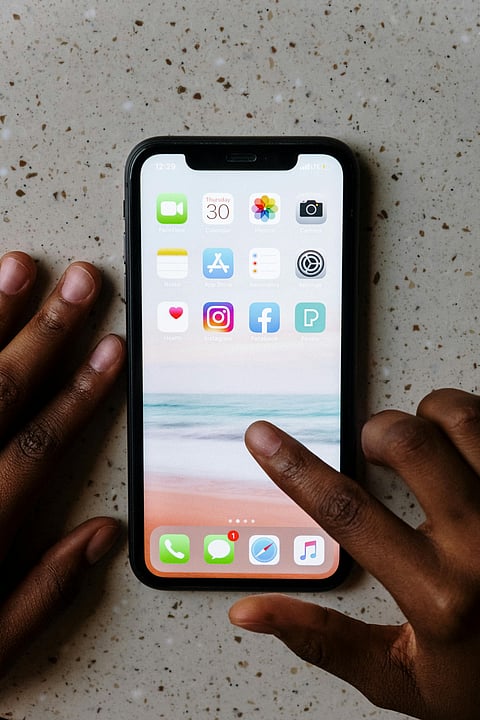 An overhead shot of a person with dark skin interacting with an iPhone resting on a light-speckled countertop. Their left hand rests beside the phone while their right index finger touches the screen, which displays various app icons like Instagram and Facebook over a beach wallpaper.