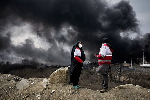 Two women from the Iranian Red Crescent Society stand as a thick plume of smoke from a U.S.-Israeli strike on an oil storage facility late Saturday rises in the sky in Tehran, Iran, Sunday, March 8, 2026.