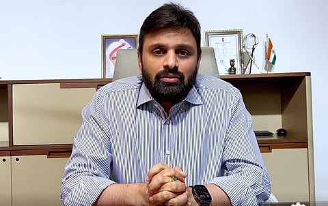 A medium shot of a man with a dark beard and mustache, wearing a blue and white pinstriped button-down shirt. He is sitting at a desk with his hands clasped, looking directly at the camera with a serious expression. Behind him is a modern office setting featuring light-colored wooden cabinets, framed pictures, an Indian flag, and a gold trophy.