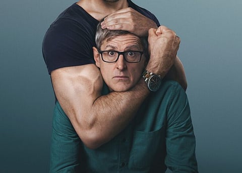 Promotional image of Louis Theroux wearing glasses and a teal shirt, looking at the camera, while a muscular man standing behind him wraps one arm around his neck and places a hand on his head against a plain blue-grey background.