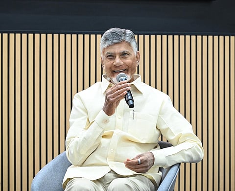 A medium shot of N. Chandrababu Naidu, the former Chief Minister of Andhra Pradesh, sitting in a modern grey chair. He is smiling warmly while holding a microphone to his mouth as if speaking at an event.

He is dressed in his signature light-cream, short-sleeved button-down shirt and matching trousers. The background features a minimalist wall with vertical light-wood slats over a dark panel. The lighting is bright and professional, highlighting his grey hair and expressive gestures.