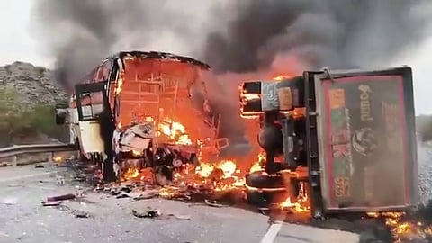 A high-angle, eye-level, medium shot shows a bus and a truck on fire on a highway. The bus is on the left, and the truck is on the right. Both vehicles are engulfed in orange and yellow flames, and thick black and grey smoke rises from them. The bus's interior is visible through the windows, and it is filled with fire. The truck's cabin is also on fire. Debris from both vehicles is scattered on the highway. In the background, there are hills and a cloudy sky. The lighting is dim, suggesting it is either dawn or dusk.