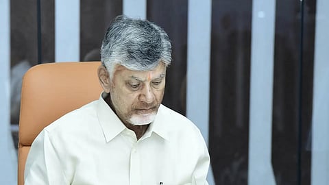 The image shows Nara Chandrababu Naidu, an Indian politician and leader of the Telugu Desam Party (TDP).

He is seated at a white desk in a professional office setting, wearing a light-colored, button-down shirt. He has grey hair and a white beard, and a small orange 'tilak' is visible on his forehead. Naidu is focused on a document in front of him, turning a page with his right hand. The background features a modern, striped wall pattern and a tan-colored office chair.