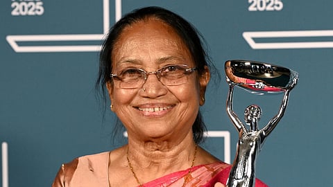 An elderly woman wearing glasses and a pink sari smiles while holding a silver trophy against a teal backdrop with “2025” visible, at the International Booker Prize event.