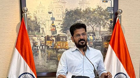 Revanth Reddy stands at a black podium centered between two Indian flags. He is wearing a light-colored, long-sleeved button-down shirt and glasses. The podium features the official seal of the Government of Telangana. Behind him is a large painting depicting a busy street scene in front of the Charminar in Hyderabad.