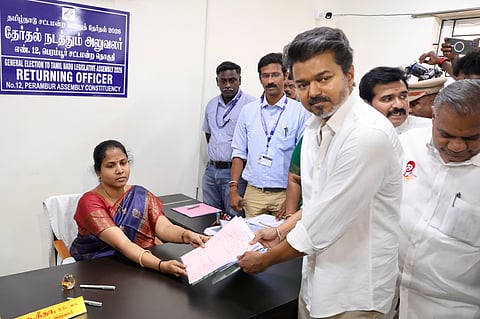 A high-angle, medium shot shows Tamil actor and politician Vijay, the chief of the Tamilaga Vettri Kazhagam (TVK) party, submitting his nomination papers for the 2026 Tamil Nadu Legislative Assembly election.

Vijay, dressed in a crisp white shirt, stands on the right, handing over a stack of documents to a female election official seated behind a desk on the left. The official, wearing a blue and red saree, receives the papers. Behind them stands a blue sign that reads "GENERAL ELECTION TO TAMIL NADU LEGISLATIVE ASSEMBLY 2026, RETURNING OFFICER, No. 12, PERAMBUR ASSEMBLY CONSTITUENCY" in both English and Tamil. Several other men, including security or party aides, are visible in the background, observing the process.