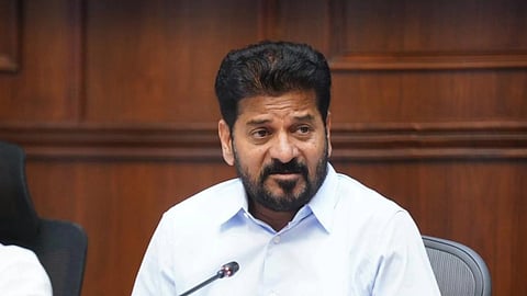 Revanth Reddy with dark, wavy hair and a groomed beard and mustache sits at a long wooden conference table. He is wearing a light blue button-down shirt and has his hands clasped in front of him. On the table before him are a desktop microphone and a box of tissues with the word "PREMIER" printed on it in gold lettering. The background consists of dark wood-paneled walls, suggesting a formal office or meeting room setting.