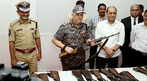 A medium shot captures a high-ranking Indian police official, identified by his name tag as Harish, in a digital camouflage uniform and cap, standing behind a white table laden with several rifles. He holds one rifle horizontally, his right hand on the pistol grip and his left hand on the handguard. To his left stands another officer in a tan police uniform and peaked cap, smiling. To Harish's right, a group of men in civilian clothes, including one in a white shirt and another in a dark suit, observe the scene. The background is a plain white wall, and a camera lens is visible in the bottom-left foreground, suggesting a press conference or official briefing.