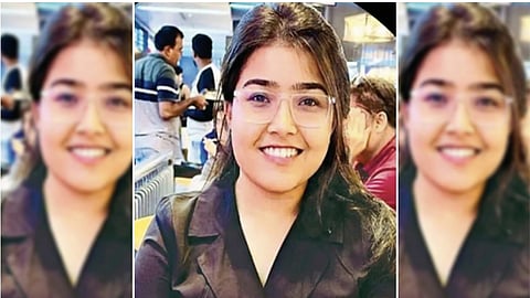 A young South Asian woman with long dark hair and clear-framed glasses smiles at the camera. She is wearing a dark blazer over a black top and is seated in what appears to be a brightly lit restaurant or cafe, with other patrons visible in the soft-focus background.