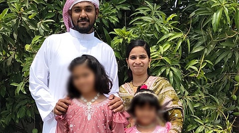 A full-length, outdoor family portrait of four people standing in front of a lush, leafy green tree.

On the left, a man with a dark beard wears a white thobe and a red-and-white checkered ghutra. Next to him, a woman with dark hair pulled back wears a green and gold patterned salwar kameez.

In front of them stand two young girls with their faces blurred for privacy. The girl on the left wears a long-sleeved, floor-length mauve dress with ruffled sleeves and a silver embellished belt. The girl on the right wears a sleeveless pink and white floral dress with a bow at the waist.

The family is standing on a light gray tiled patio under bright, natural light.