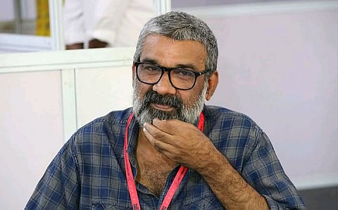 A middle-aged man with grey hair and beard, wearing glasses and a checkered shirt, sits indoors with his hand resting on his chin.