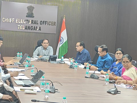 The image shows a group of officials seated around a large wooden conference table in an office. A sign on the wall behind the central figure reads "CHIEF ELECTORAL OFFICER TELANGANA" below the State Emblem of India. An Indian national flag stands to the right of the sign.

The officials, both men and women, are dressed in formal or semi-formal attire and appear to be engaged in a meeting. Several laptops, microphones, notebooks, and water bottles are spread across the table. The room has dark wood-paneled walls, creating a formal atmosphere.