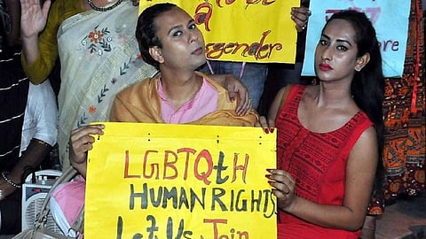 Two transgender persons stand at a protest holding a yellow placard that reads “LGBTQ+ human rights, let us join, let us fight,” while others with similar signs are visible in the background.