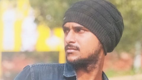 A young man with a beard and mustache looking off to the side. He is wearing a dark denim shirt and a black knit beanie. He is standing behind a yellow horizontal bar, with a blurred outdoor background.