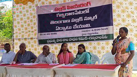 A wide-angle outdoor shot of a public hearing in Gabbilalapet, Telangana. Five people (three men and two women) are seated behind a long table covered with white and red cloths. To the right, a woman in a peach-colored floral saree stands, speaking into a microphone.

Behind the group is a large banner written in Telugu saying a Telangana Public School should be established in Gabbilalapet.

The setting is under a temporary tent structure with blue and white fabric overhead and a patterned yellow backdrop. The atmosphere appears formal and community-focused.