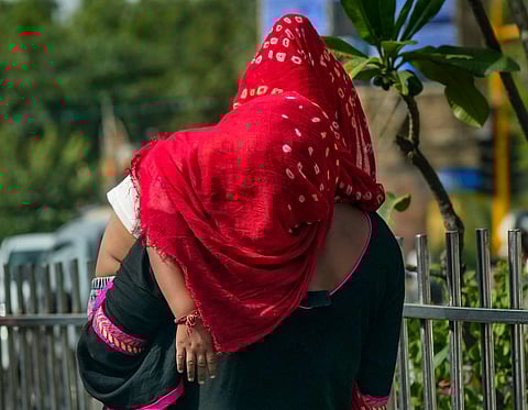 A rear view of a mother carrying a child on her back outdoors. Both are covered with a vibrant red and white patterned dupatta (scarf) to shield themselves from the sun. The mother is wearing a black garment with pink and gold accents, and they are standing near a metal railing with greenery and a blurred street in the background.
