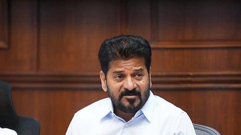 A medium shot of Anumula Revanth Reddy, the Chief Minister of Telangana, seated at a dark wooden conference table against a wood-paneled wall. He is a middle-aged man with dark hair and a groomed beard, wearing a light blue button-down shirt. His hands are clasped on the table in front of him. A black conference microphone and a tissue box with "PREMIER" printed in gold letters are positioned on the table before him. He appears to be speaking or listening intently during a meeting.