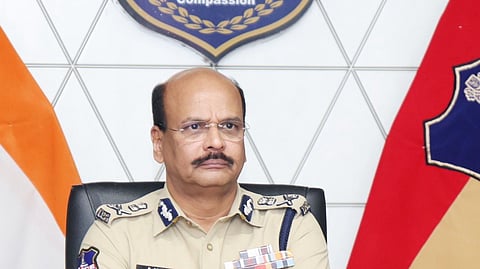 A formal portrait of B. Shivadhar Reddy, a high-ranking official of the Telangana Police, seated at a wooden desk with his arms crossed.

He is wearing a khaki police uniform featuring several service ribbons on his chest, a name tag, and shoulder insignia indicating his rank. He has short, thinning dark hair, a mustache, and wears gold-rimmed glasses and a gold watch.

In the background, a large Telangana Police emblem is mounted on a white geometric-patterned wall. The emblem includes the State Emblem of India and the motto "Duty, Honour, Compassion." To his left is the Indian National Flag, and to his right is the Telangana Police flag. A microphone, a bottle of water, and some tissues are visible on the desk in the foreground.