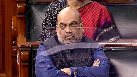 A medium shot of Amit Shah, the Union Home Minister of India, seated in Parliament. He is bald with a grey beard and mustache, wearing gold-rimmed glasses, a blue long-sleeved shirt, and a dark grey Nehru jacket. He sits with his arms crossed, looking toward the left with a serious expression. Behind him, a person in a red and black patterned sari is partially visible. The setting features ornate wooden furniture and a glass partition in the foreground.