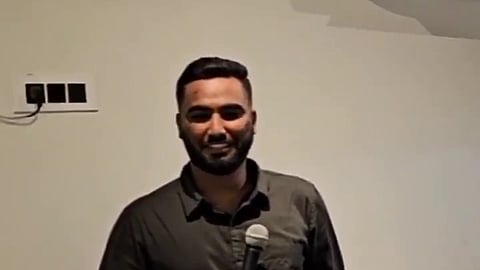 A man with a beard and a bandage on his right hand stands on a small stage, smiling while holding a microphone. He wears a dark, long-sleeved button-down shirt and light-colored pants. In the foreground, the back of a person’s head with dark hair in a ponytail is visible, and a tall black stool stands to the man's right. The background features a plain white wall with electrical outlets and an air conditioning unit.