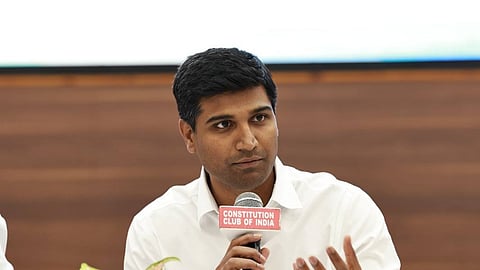 A medium shot of Lavu Sri Krishna Devarayalu, an Indian politician, seated at a table and speaking into a microphone. He is a young man with short dark hair, wearing a crisp white button-down shirt. His right hand holds a microphone labeled "CONSTITUTION CLUB OF INDIA," while his left hand is raised in a gesture to emphasize his point.
