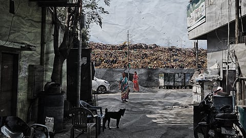 A narrow alley in an urban area opens onto a street where two women in colorful saris walk past a large landfill piled high with trash in the background. A stray dog stands in the foreground near plastic chairs and scattered debris, with buildings, bins, and a parked car framing the scene.