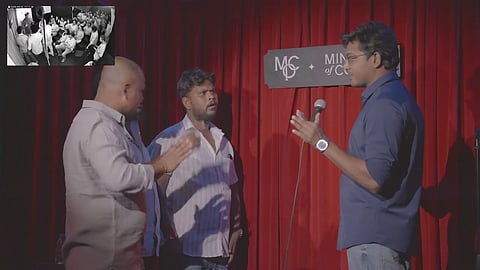A medium shot captures a stand-up comedian on a stage with a red curtain backdrop, interacting with two audience members. The comedian, a South Asian man in a dark blue button-down shirt and glasses, holds a microphone and gestures toward the two men standing in front of him. One man is bald and wears a light-colored shirt, while the other has dark hair and a white collared shirt; both appear to be engaged in a tense or serious conversation with the performer. In the top left corner, an inset black-and-white security camera feed shows a wider view of the dimly lit comedy club and the seated audience. A sign on the curtain behind the comedian reads "MOC + Ministry of Comedy."