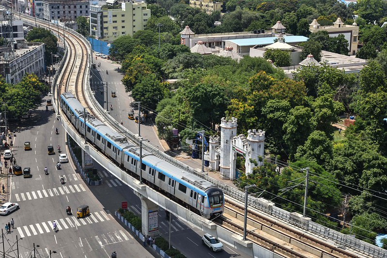 An aerial view shows a blue and silver Hyderabad Metro train traveling on an elevated track that curves through the city. Below the tracks is a multi-lane road with several white cars, yellow and black auto-rickshaws, and motorcycles. To the right of the tracks, there is a large area of lush green trees, behind which several buildings with traditional Indian architectural features and white domes are visible. A white, castle-like gate stands near the edge of the trees next to the road. The sky is bright, and the scene is captured in clear daylight.