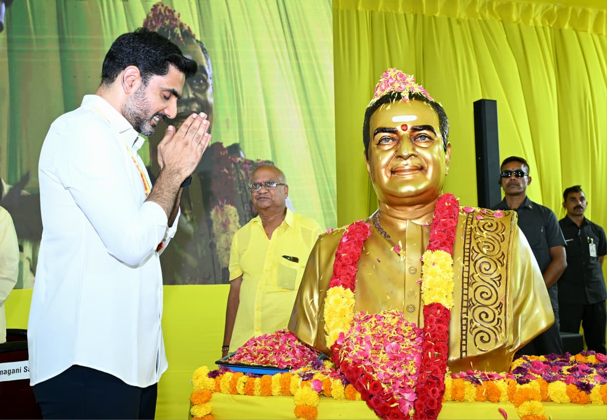 Nara Lokesh with a beard, wearing a white shirt, stands with his hands pressed together in a respectful namaste gesture before a golden bust of N.T. Rama Rao. The bust is adorned with red and yellow flower garlands. In the background, another man in a yellow shirt looks on, and security personnel stand behind the statue against a bright yellow backdrop.