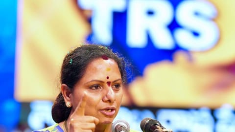 A medium shot of Kalvakuntla Kavitha, an Indian politician, speaking at a podium. She is wearing a lime green saree with a blue border and has her right hand raised with her index finger pointed upward as if making a strong point. Behind her, a large digital screen displays the letters "TRS" in white over a blue map of Telangana against a yellow background. The lighting is bright and stage-focused, emphasizing her animated expression.