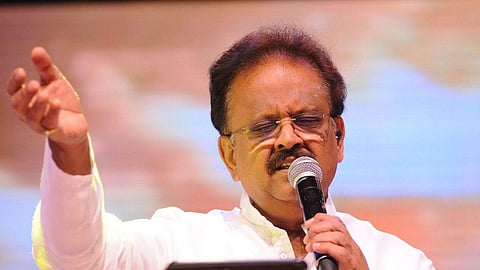A close-up photograph of S. P. Balasubrahmanyam, a renowned Indian playback singer, performing on stage. He is wearing a white shirt, has a mustache and is wearing glasses. He holds a microphone in his left hand near his mouth, and his right hand is raised slightly to the side in a gesture. He appears to be singing with his eyes closed or looking down. The background is brightly lit with warm, out-of-focus lights.