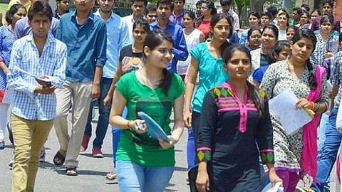 A large group of college-aged students, both men and women, are walking together outdoors in bright daylight. Most are casually dressed, holding papers or folders, suggesting they may be coming from or going to an exam or class. The atmosphere appears busy and energetic, with trees and a building visible in the background.