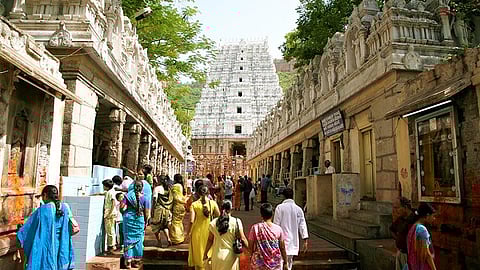 People walk towards a large, intricately carved temple in Tirumala in an outdoor setting, surrounded by trees and detailed stone architecture
